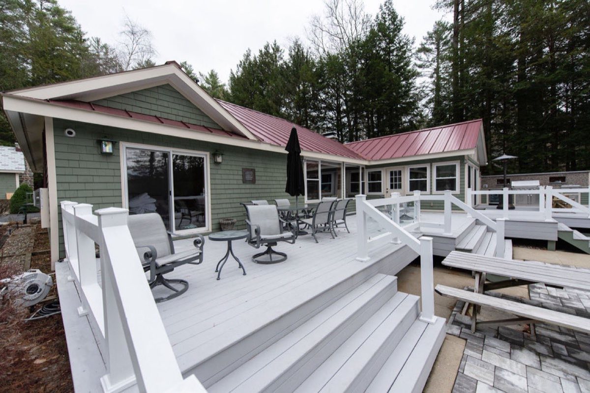 The waterfront deck at Camp Sunnyside set with dining chairs and a table, the sage-shingle house and lake behind.