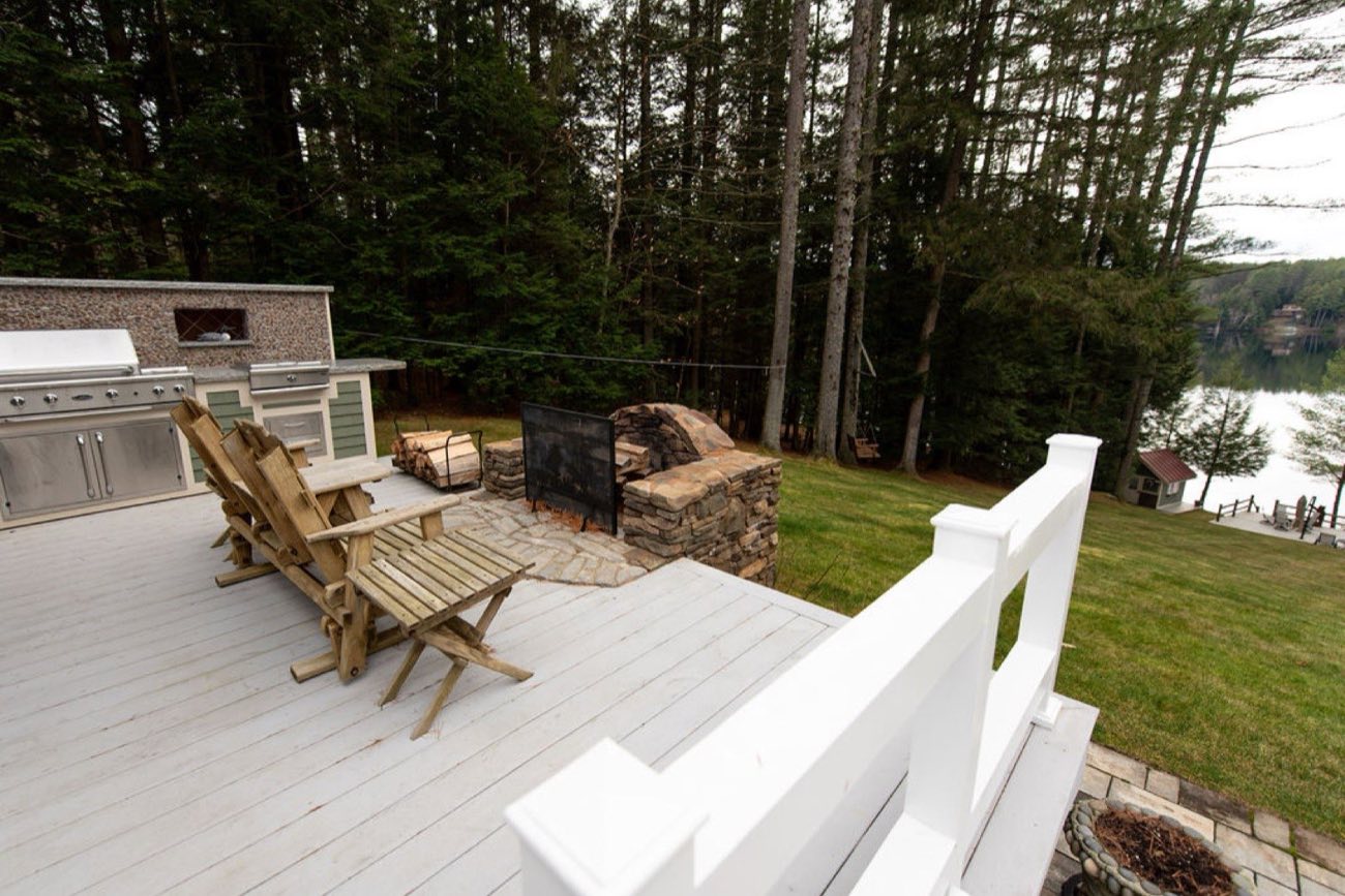 The fieldstone outdoor grill, Adirondack chairs, and fire area on the deck at Camp Sunnyside with the lake beyond the pines.