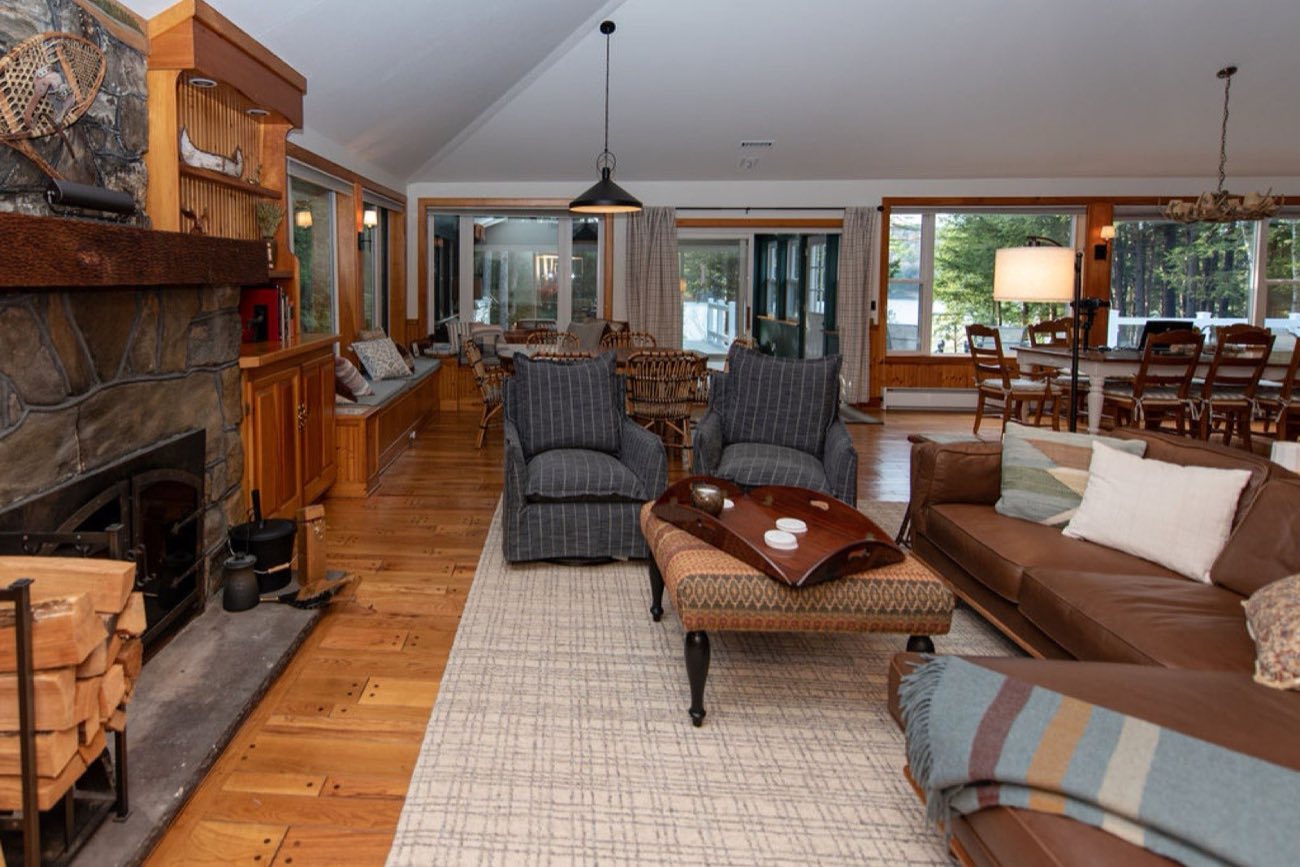 The great room at Camp Sunnyside’s main house with a fieldstone fireplace, snowshoes and a canoe paddle over the mantel, honey-pine floors, and a wall of glass to the pines.